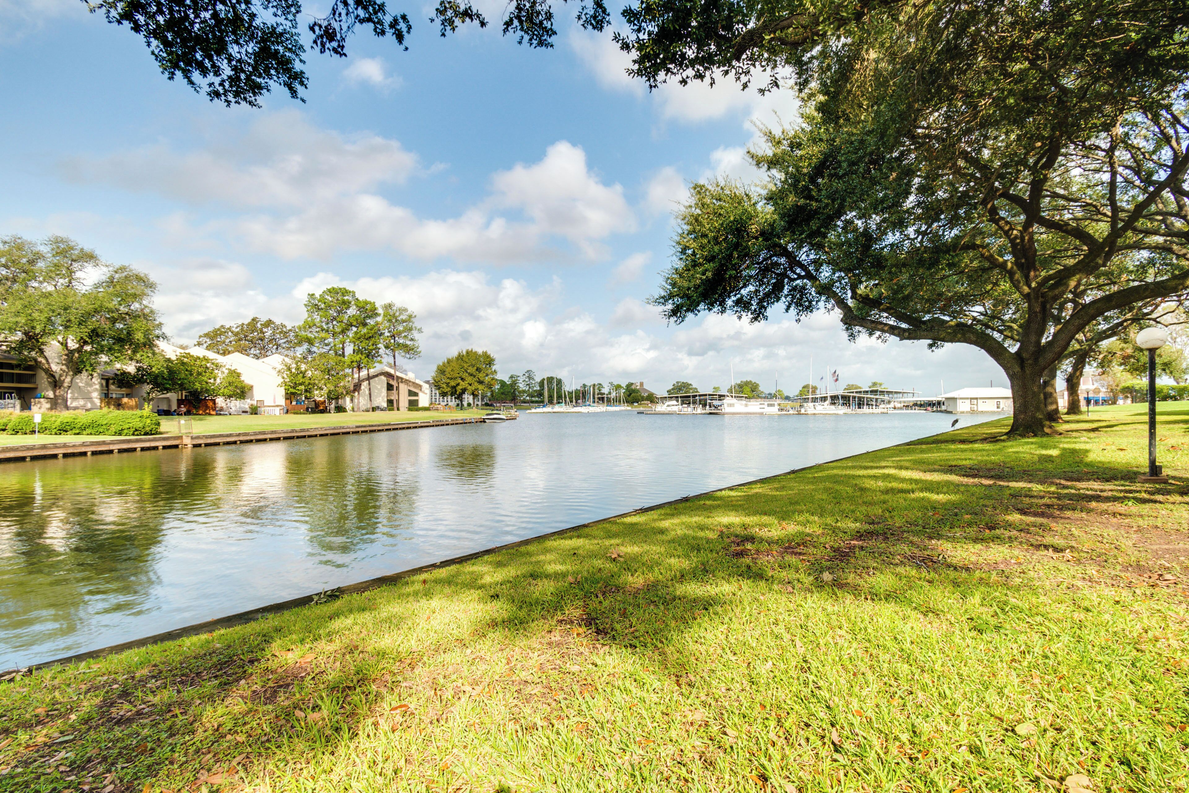 A calm waterway with a grassy bank, trees, and buildings in the background.