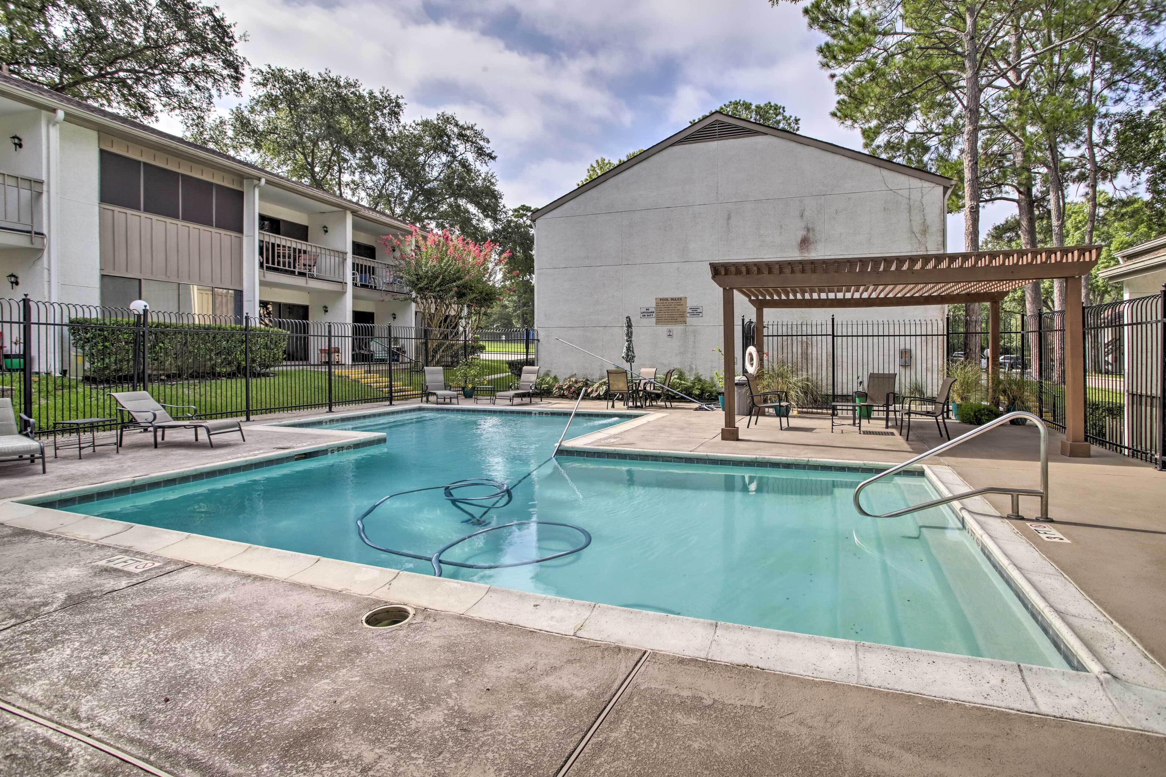 A rectangular pool with a pergola and lounge chairs, surrounded by a fence and a building with a balcony.