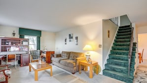 Ein Wohnzimmer mit Sofa, Couchtisch und Schreibtisch mit Computer. Eine Treppe mit grünem Teppich führt in einen anderen Raum.