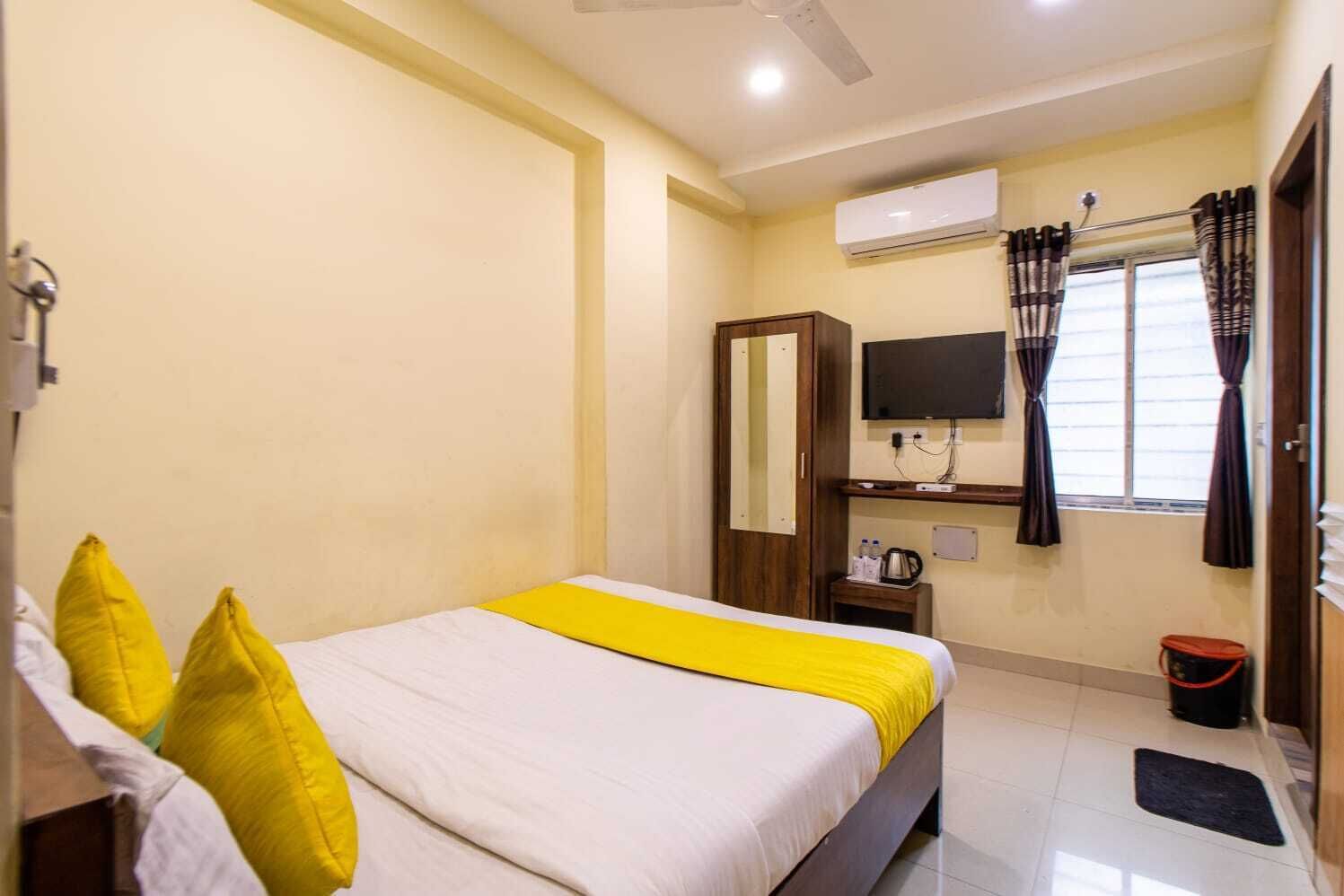 A hotel room with a bed, a TV, an air conditioning unit, a window with curtains, and a small table with a kettle.