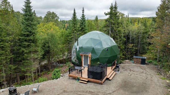 Een geodetische koepelstructuur met een houten terras, een kleine deur en een zithoek met stoelen.