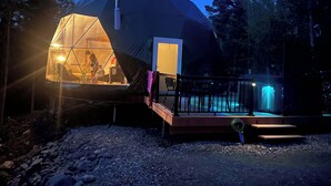 Une structure en forme de dôme géodésique dotée d’une entrée en verre, d’une terrasse et d’un petit espace extérieur éclairé d’une lumière bleue.