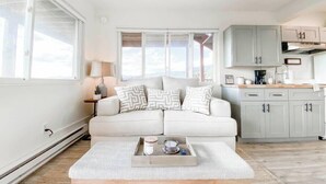 A modern living room with a white sofa, patterned cushions, a coffee table with a tray, and a kitchen area with light-colored cabinets and a view through the windows. - Scenic Mountainside Studio (Steamboat Springs)