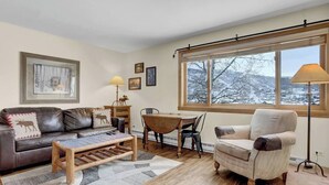A cozy living room with a brown leather sofa, a wooden coffee table, a beige armchair, and a dining table with chairs. There are framed pictures on the wall and a window with a view of a snowy landscape. - Gondola Village Chalet (Steamboat Springs)