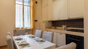 Ein Essbereich mit einem weißen Tisch, gedeckt für vier Personen, eine Küche mit weißen Schränken und einer schwarzen, gefliesten Rückwand sowie ein Fenster mit gelben Vorhängen.