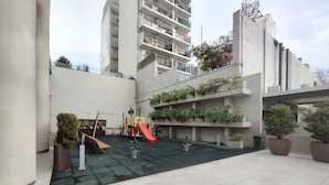 Une cour extérieure avec un espace de jeux pour enfants, des plantes en pot et un bâtiment en arrière-plan.