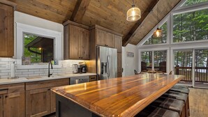 A modern kitchen with wooden cabinets, a large island with bar stools, a stainless steel refrigerator, and a window overlooking a forest. - Gorgeous Lakehouse, Sandy Bottom, Dock, AC (Lac du Flambeau)