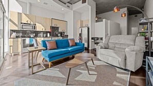 Ein modernes Wohnzimmer mit einem blauen Sofa, einem grauen Sessel und einem Couchtisch. Im Hintergrund ist eine Küche mit Mikrowelle, Herd und Kühlschrank zu sehen.
