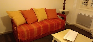 Ein rotes Sofa mit gestreifter Bettwäsche und vielen Kissen, ein kleiner weißer Tisch mit einer Spitzenleinentischdecke und ein weißer Heizkörper in einem Raum mit schlichten Wänden.
