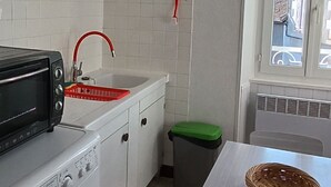 Pieni, käytännöllinen keittiö, jossa on pyykinpesukone, leivänpaahdin, punaisella hanalla varustettu pesuallas ja seinällä oleva kello.