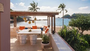 Un área exterior con mesas y sillas, una pérgola y vistas a la playa y al océano.