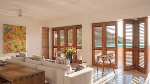 Una sala de estar moderna con un sofá, una mesa de madera y vista al mar a través de amplios ventanales con contraventanas de madera.