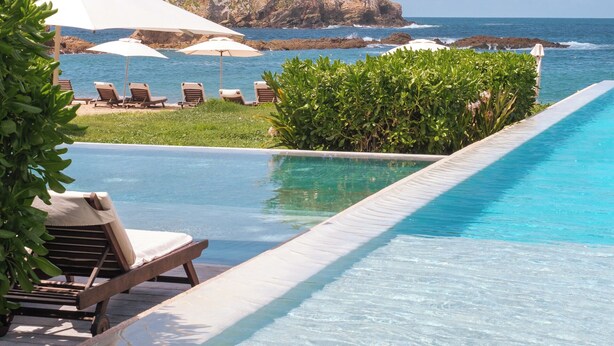 Zona de piscina con sillones de descanso y sombrillas, con vista despejada al océano y una isla rocosa al fondo.
