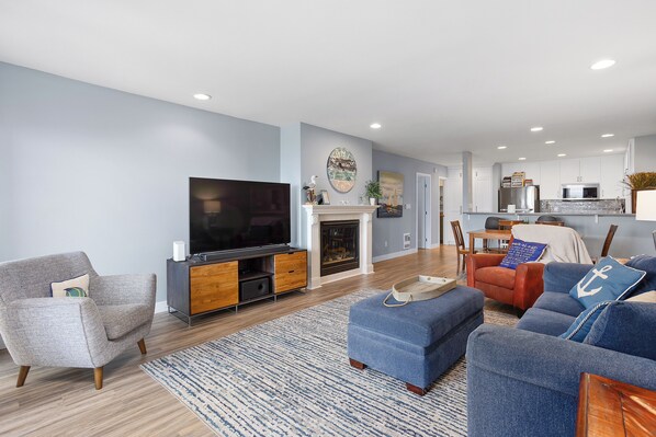 A modern living room with a flat-screen TV, a fireplace, and a dining area with a kitchen in the background.