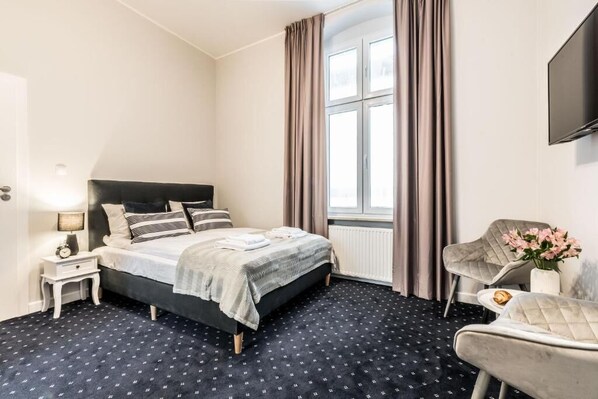 A hotel room with a bed, a nightstand, a chair, a table with a flower vase, and a TV mounted on the wall. - SERENITY Residence (Poznan)