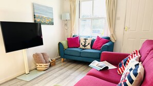 A living room with a blue sofa, green and red armchairs, a flat-screen TV, a window with curtains, and a wall-mounted painting. - Pippin Cottage in Salcombe (Kingsbridge)