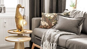 Un salon moderne avec un canapé gris, des coussins décoratifs et une sculpture d’oiseau dorée originale posée sur une petite table ronde.