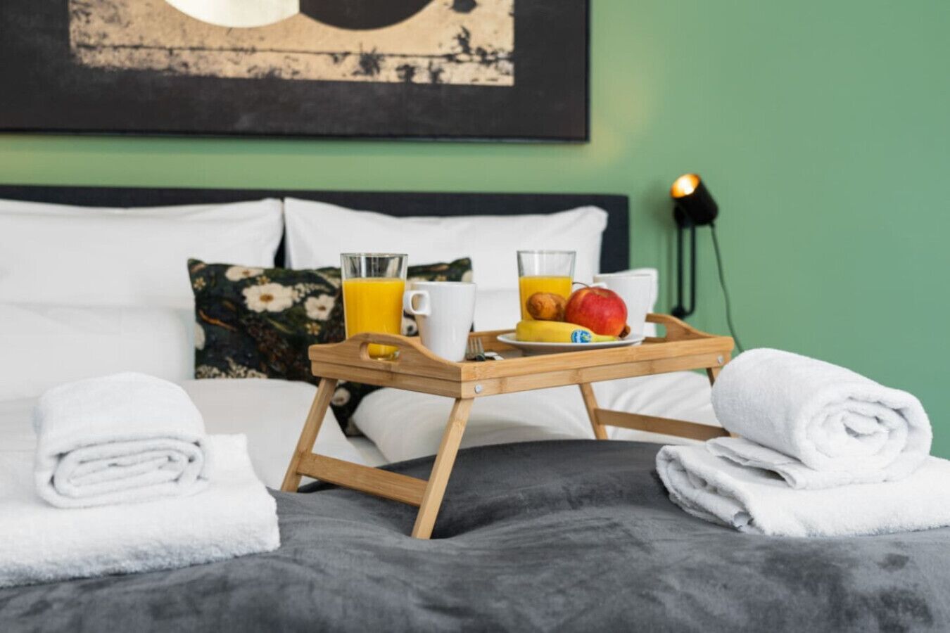 Una cama bien hecha con una bandeja de madera que contiene alimentos para el desayuno, un cojín con estampado floral y un cuadro en la pared.