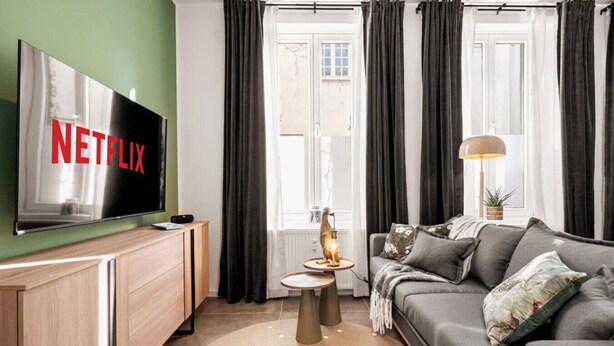 Un salon moderne avec un téléviseur à écran plat, un canapé gris, une petite table ronde avec une lampe et une grande fenêtre avec des rideaux.