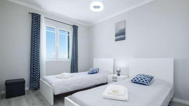 Ein modernes Schlafzimmer mit zwei Betten, einem Nachttisch mit Lampe, einer kleinen Bank und einem Fenster mit gemusterten Vorhängen.