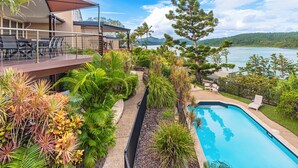 Zona de piscina con vistas a un lago, rodeada de plantas tropicales y una terraza con mobiliario exterior.
