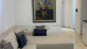 A living room with a beige sofa, a framed floral painting, and a hallway leading to a door. - Serenity Home (La Marsa)