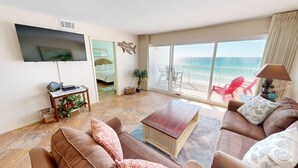 Un salón con un amplio ventanal con vistas al mar, un televisor de pantalla plana montado en la pared y un balcón con sillas rojas.
