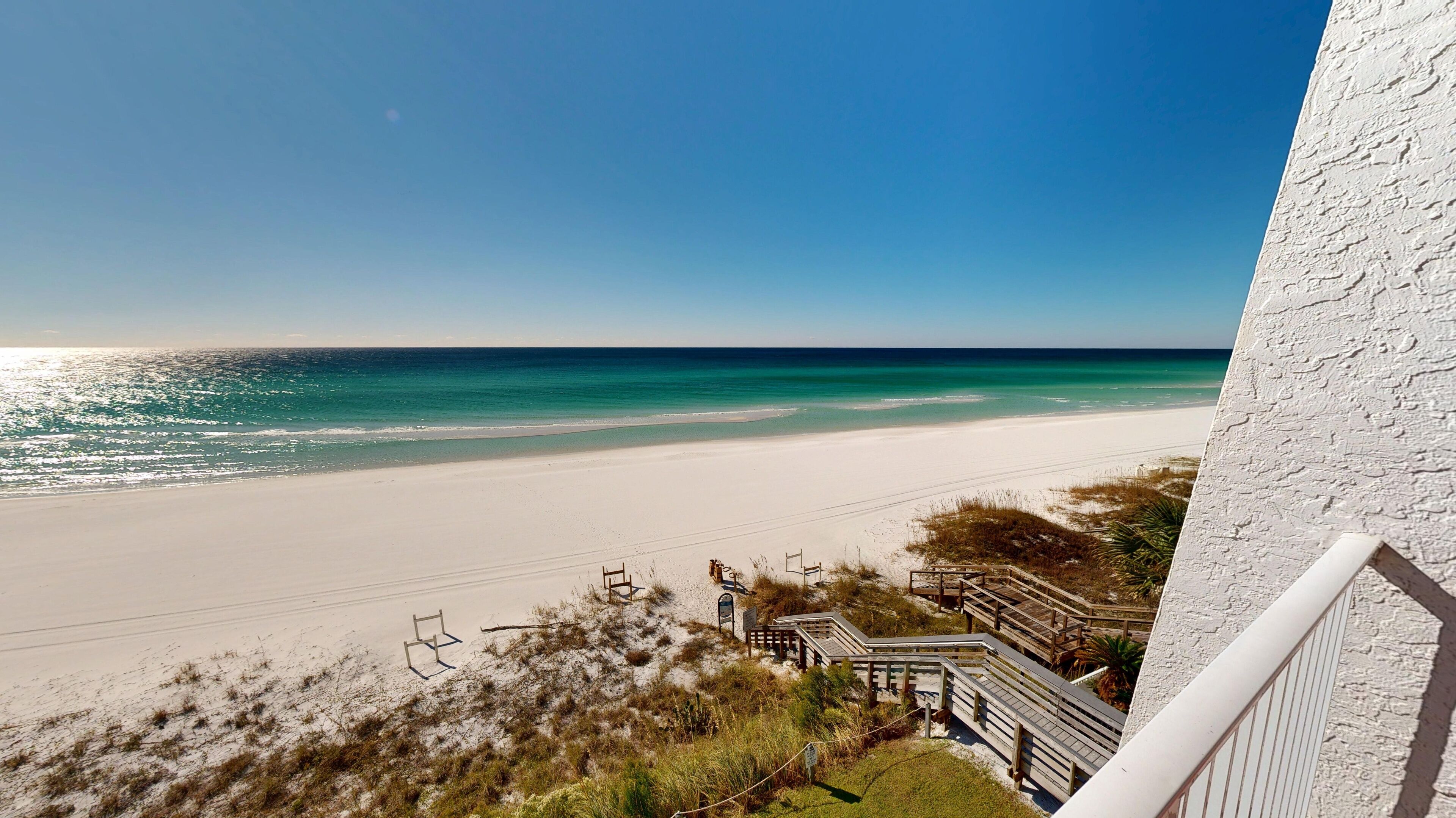 A beach view from a balcony with a clear sky, sandy beach, and wooden walkways.