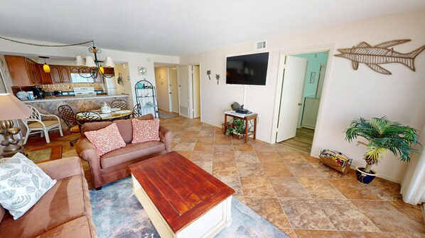 A living room with a brown sofa, a wooden coffee table, and a TV mounted on the wall. There is a dining area with a table and chairs, and a kitchen in the background. - Beach House 401d 1 Bedroom Condo (Miramar Beach)