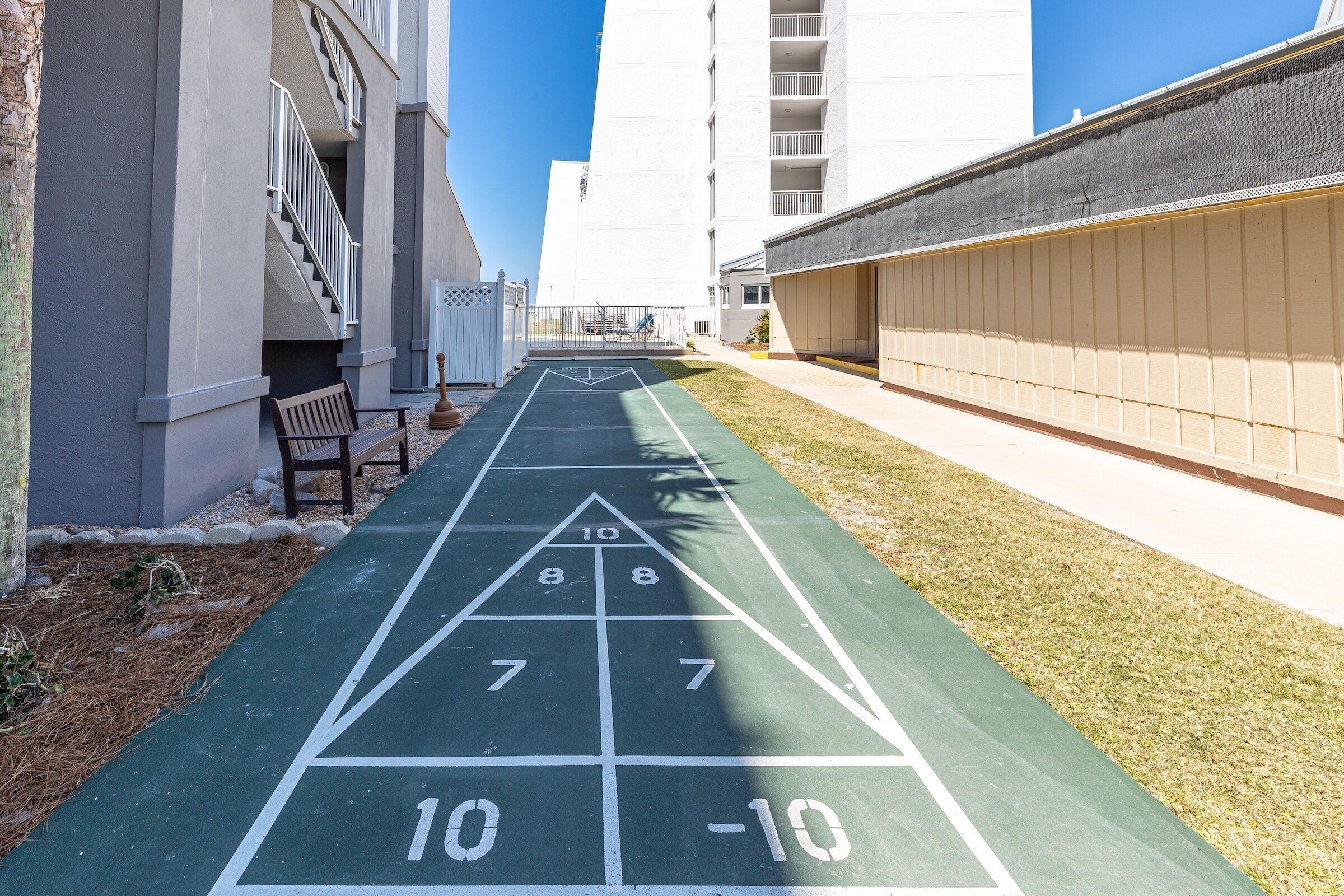 Una cancha de shuffleboard verde con números y flechas, bordeada por edificios y un banco.