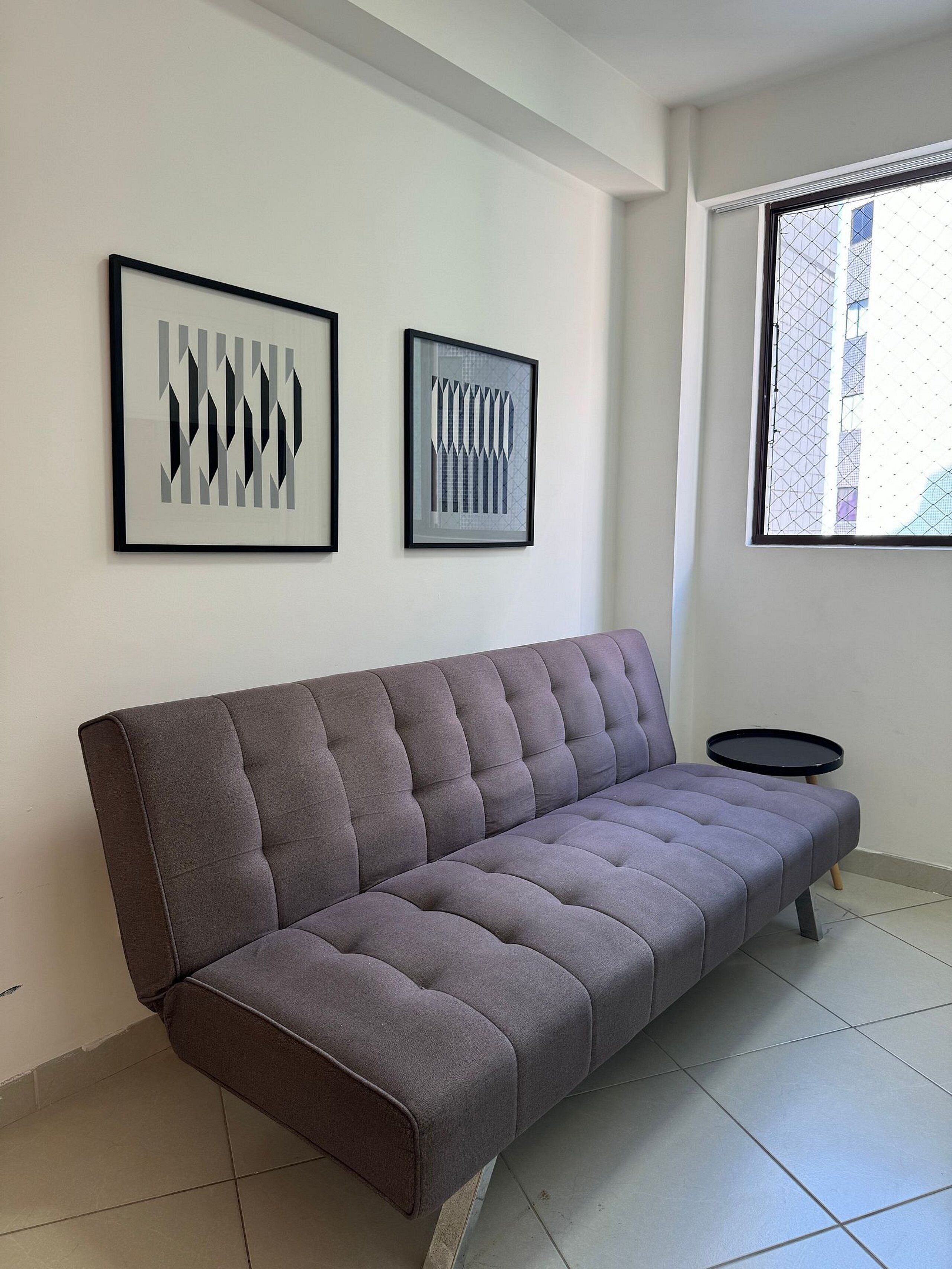 A modern living room with a grey sofa, two framed abstract artworks, a small black side table, and a window with a view of buildings.