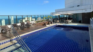 Uma piscina no terraço com azulejos azuis, espreguiçadeiras e vista para o oceano.