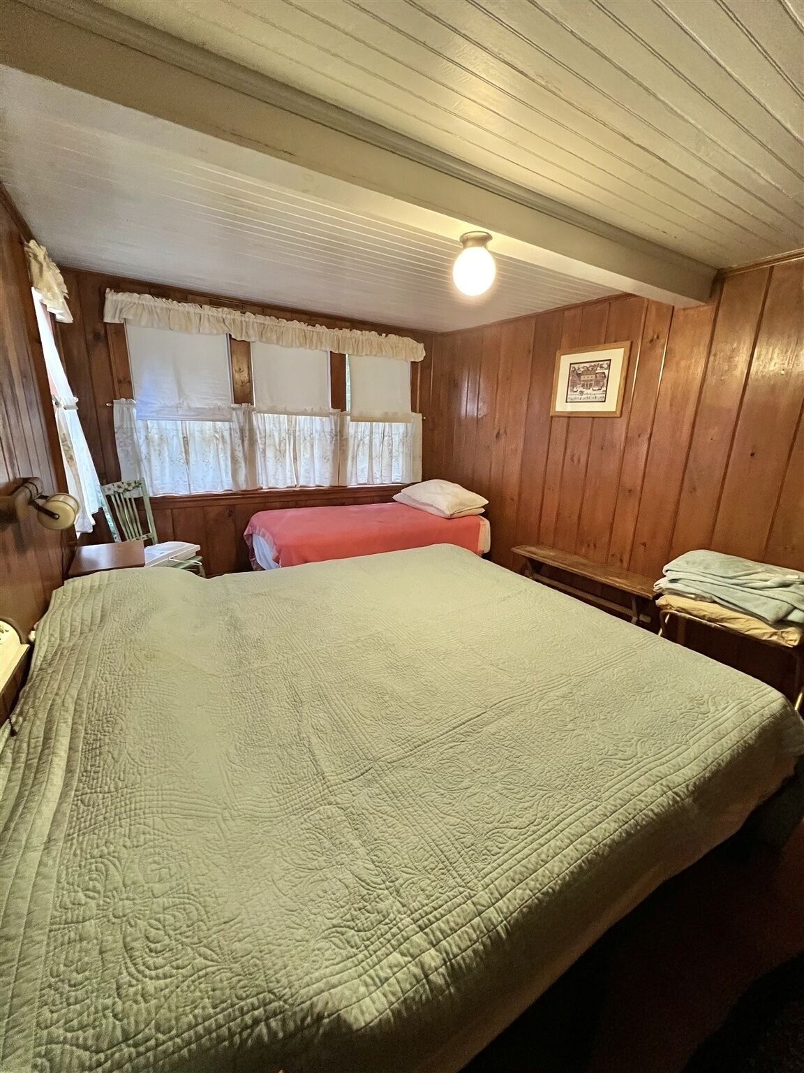 A room with a large bed, a smaller bed, a bench, and a framed picture on the wall.