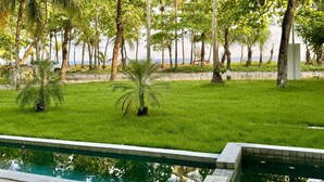 A poolside view with a grassy area, palm trees, and a beach in the background. - Esterillos Lote 6 Bedroom Home (Parrita)