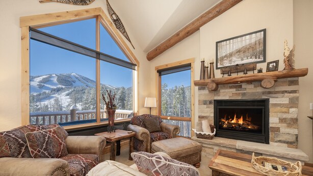 A cozy living room with a fireplace, large windows with a mountain view, and rustic decor. - Antler At Lakota 407 3 Bedroom Home (Winter Park)