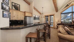 A kitchen with wooden cabinets, a granite countertop, and a bar stool. There is a couch with pillows and a view of a snowy landscape through a large window. - Antler At Lakota 407 3 Bedroom Home (Winter Park)