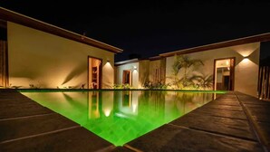 Yökuva uima-allasalueesta, jossa vesi on kirkasta. Allasta ympäröivät rakennukset, joiden ovet ovat auki ja joista loistaa lämmin valo.