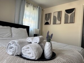 A neatly made bed with rolled white towels and a vase with lavender flowers on a tray.