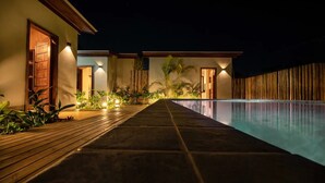 A nighttime view of a resort with a swimming pool, wooden deck, and well-lit doors.