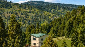 A two-story house with a green roof, surrounded by dense forest and hills. - Quinn Creek Chalet-Great home to relax with family & enjoy the views! (Bozeman)