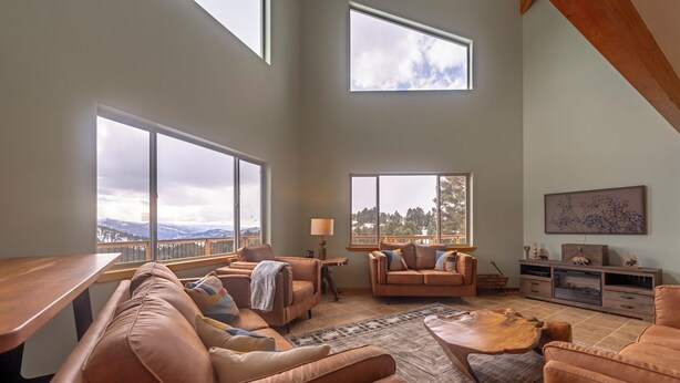 A spacious living room with large windows, a wooden table, leather sofas, and a TV stand. - Quinn Creek Chalet-Great home to relax with family & enjoy the views! (Bozeman)