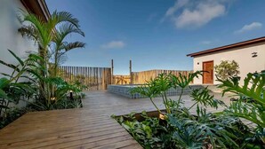 A wooden deck with tropical plants, a wooden fence, and a building with a tiled patio.