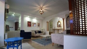 Un salon spacieux avec un plafond haut, un lustre et une cheminée. On y trouve des canapés, une table basse et un fauteuil bleu.