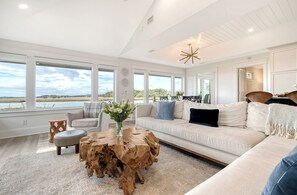 A spacious living room with a large window, a round wooden coffee table, a beige sofa, and a view of the water.