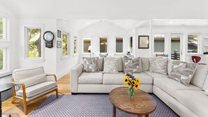 A spacious living room with a large sectional sofa, a round coffee table with a vase of flowers, and a clock on the wall. - Yards to the beach, stunning new reno (Kiawah Island)