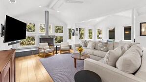 A spacious living room with a large flat-screen TV, comfortable seating, a fireplace, and large windows offering a view of greenery. - Yards to the beach, stunning new reno (Kiawah Island)