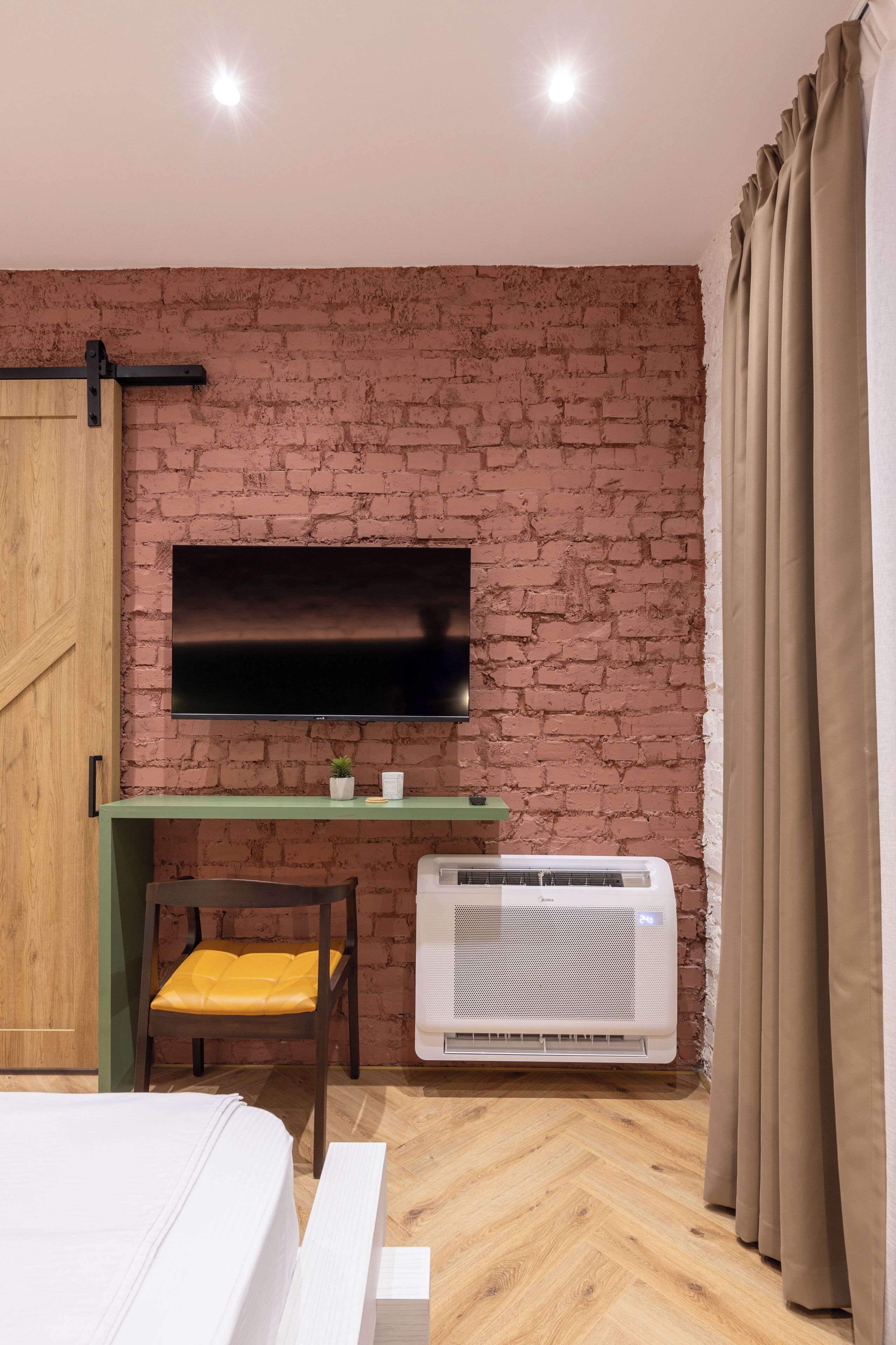 Habitación con pared de ladrillo, televisor de pantalla plana sobre un escritorio, una silla, aire acondicionado y una cama.