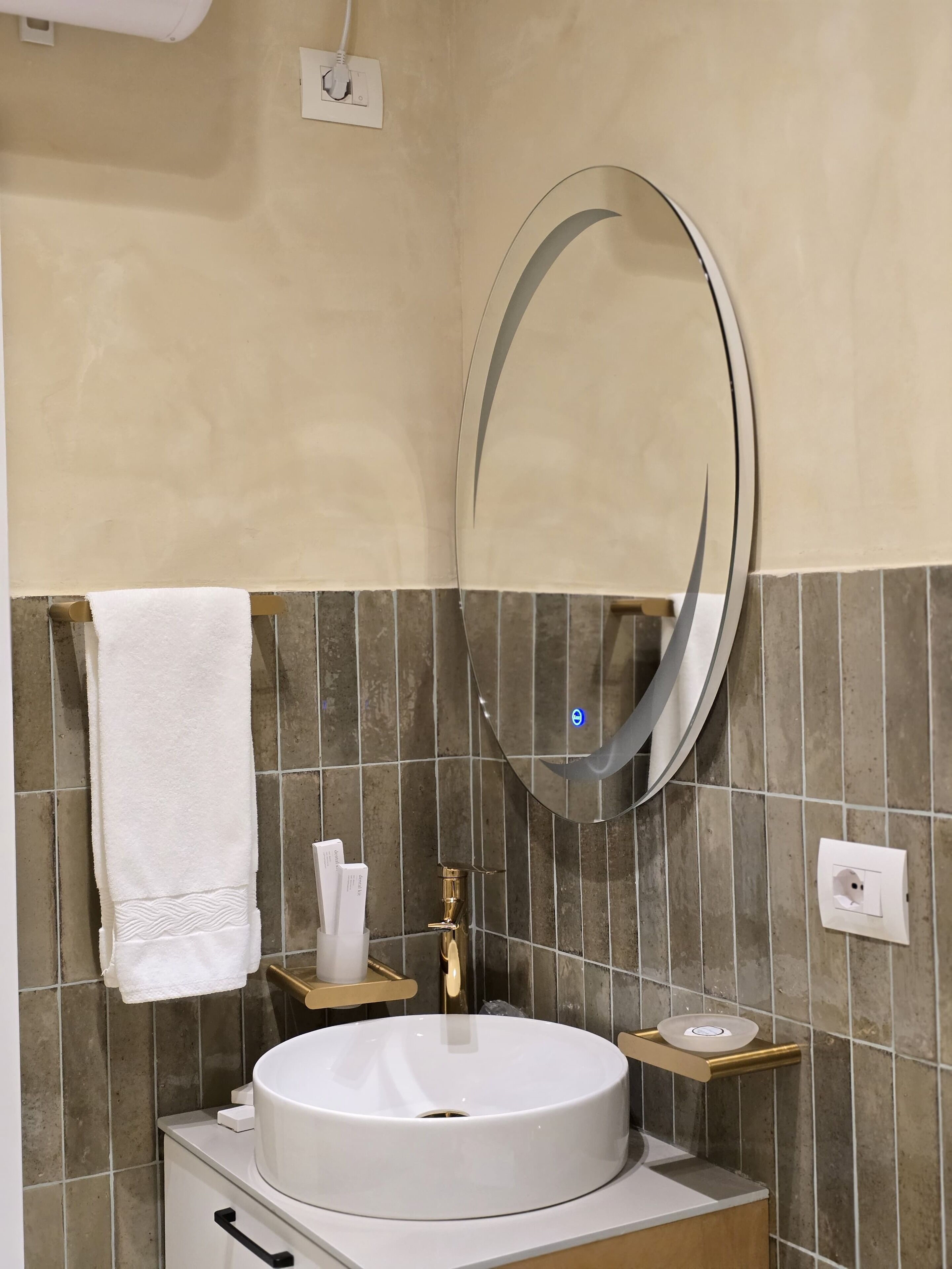 A bathroom with a round sink, a mirror, a towel, a soap dispenser, and a small shelf.
