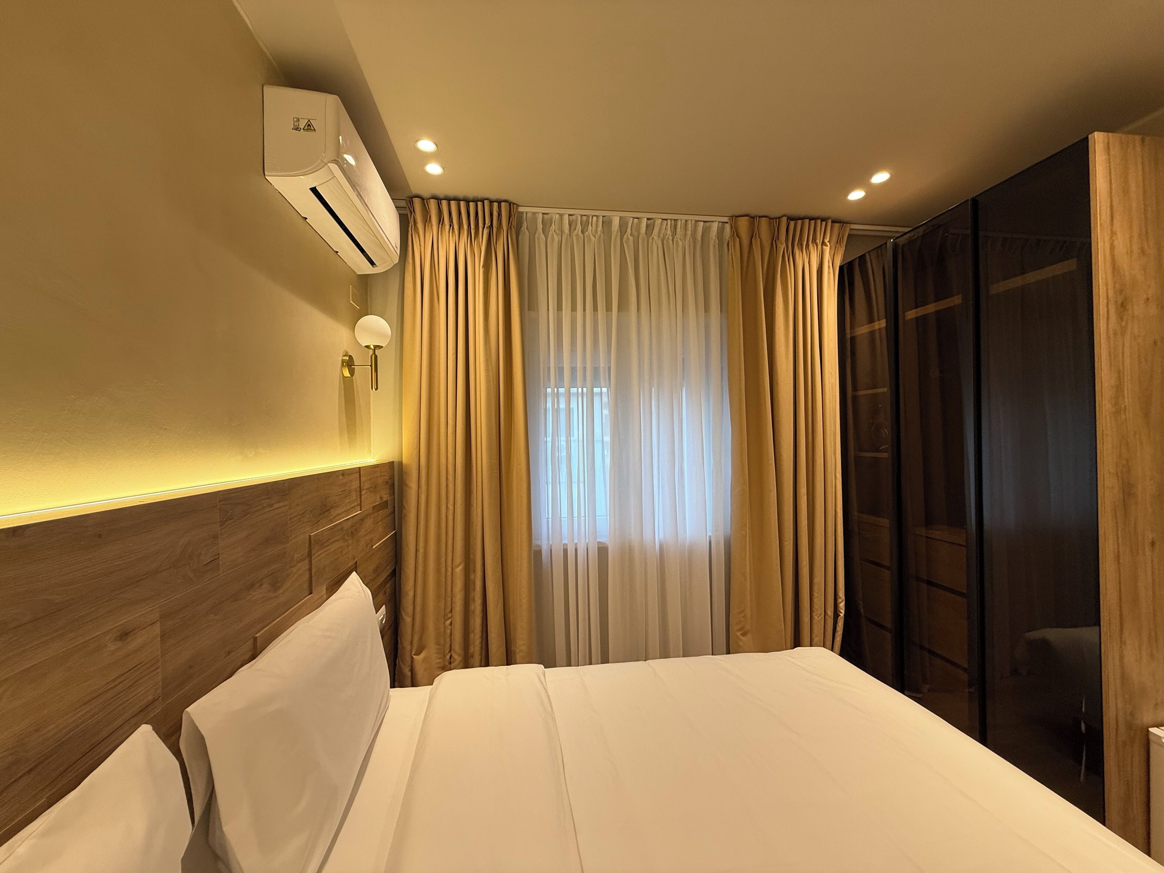 A hotel room with a bed, wooden headboard, an air conditioning unit, a wall-mounted light fixture, and a window with curtains.