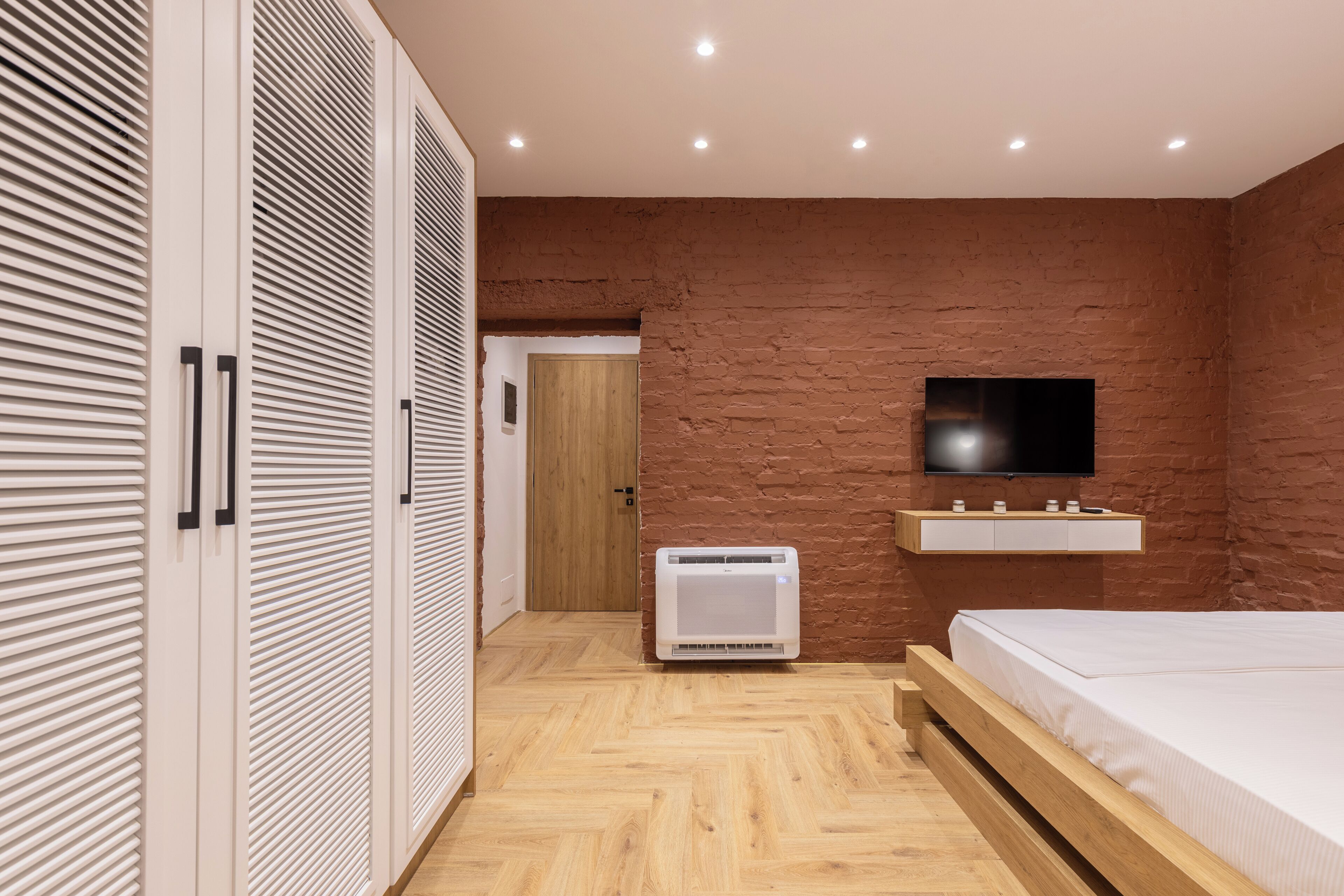 A bedroom with a wooden bed, a TV mounted on a brick wall, and a wardrobe with horizontal slats.